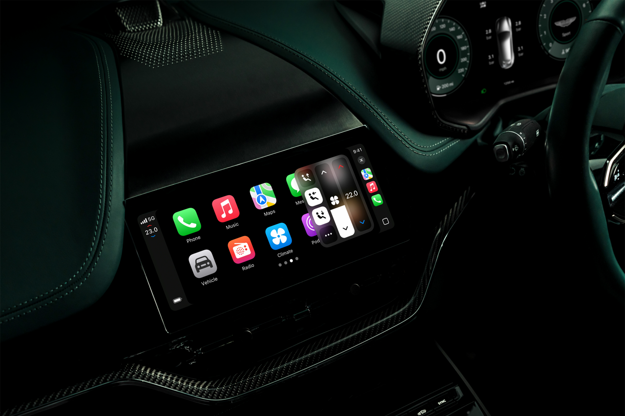 Infotainment-Display eines Aston Martin mit Apple CarPlay, Navigation, Musik, Telefon, Fahrzeug- und Klimasteuerung, 5G-Verbindung und digitalem Tachodisplay im Hintergrund.