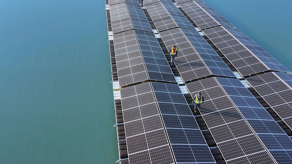 Luftaufnahme einer schwimmenden Solaranlage auf einem See. Zwei Arbeiter in Warnwesten gehen über die dicht aneinandergereihten Solarpaneele, die sich in langen Bahnen über das grünlich schimmernde Wasser erstrecken.