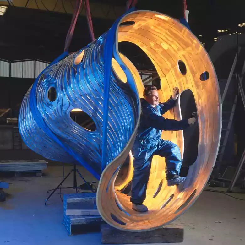 Arbeiter in blauer Schutzkleidung montiert ein großes, leuchtendes, organisch geformtes Metallbauteil in einer Werkhalle.