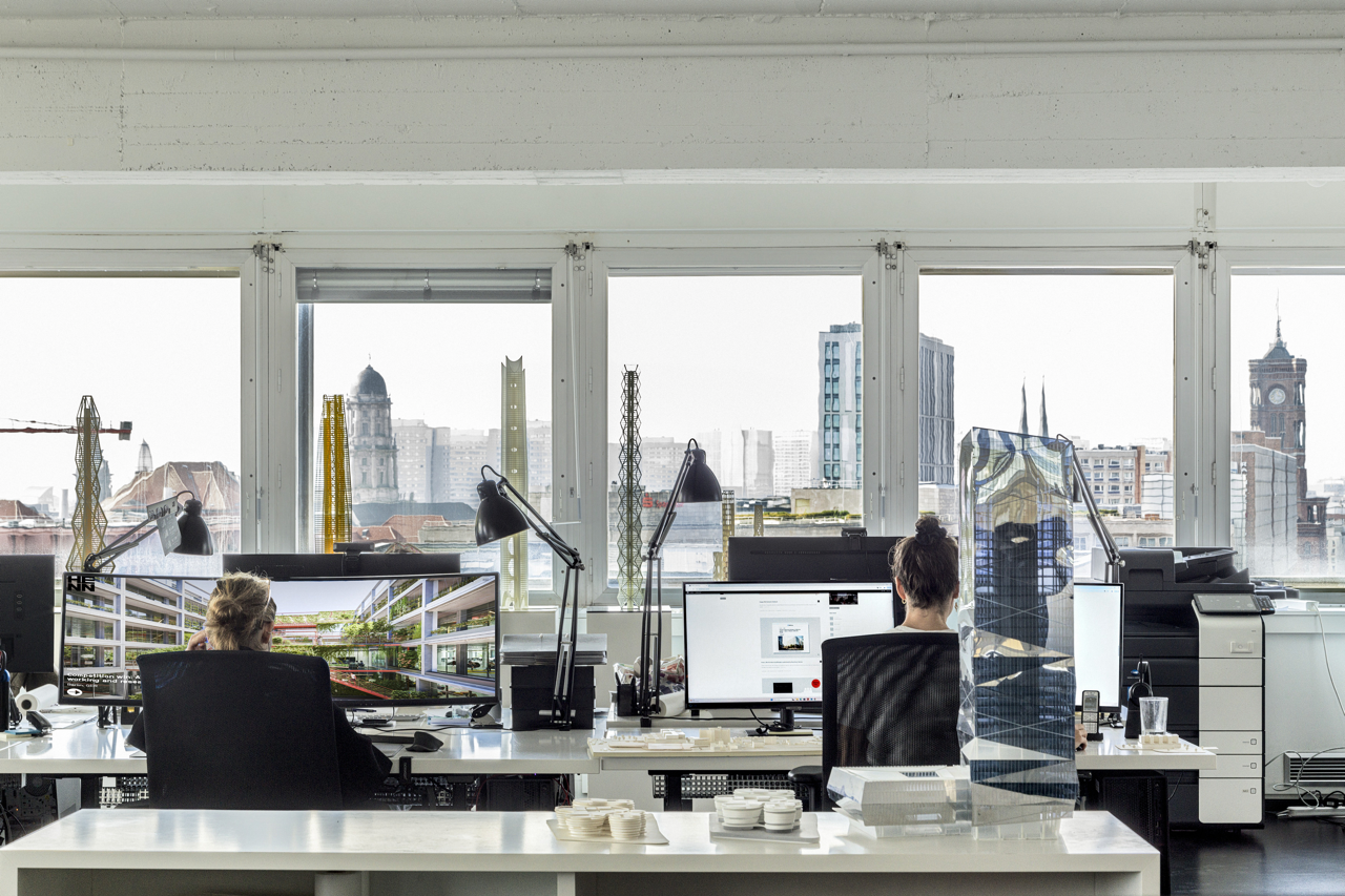 Modernes Architekturbüro in Berlin mit zwei Personen an Schreibtischen vor großen Fenstern und Blick auf die Stadt, darunter den Turm des Roten Rathauses.