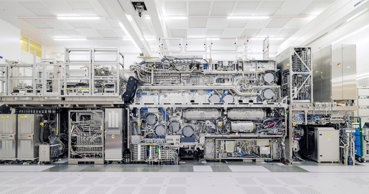 Innenansicht eines Reinraums mit einer großen EUV-Lithografiemaschine von ASML zur Chipfertigung. Die aufgeschnittene Anlage zeigt ein komplexes Geflecht aus Rohren, Kabeln, Modulen und metallischen Bauteilen unter heller Deckenbeleuchtung.