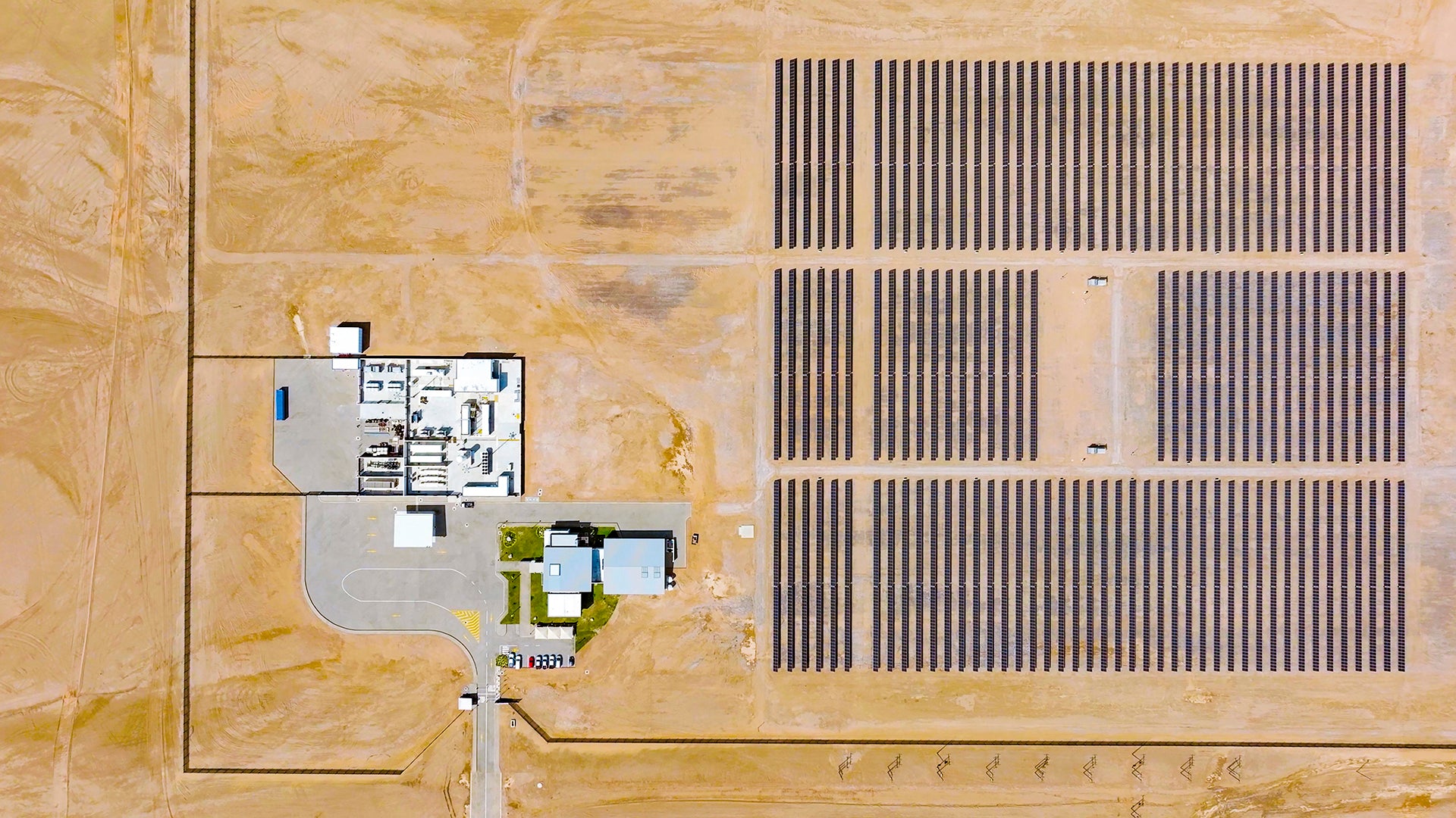 Luftaufnahme eines großen Solarfelds in einer Wüstenlandschaft mit rechteckigen Reihen von Photovoltaikmodulen und einem angrenzenden Betriebsgebäude.