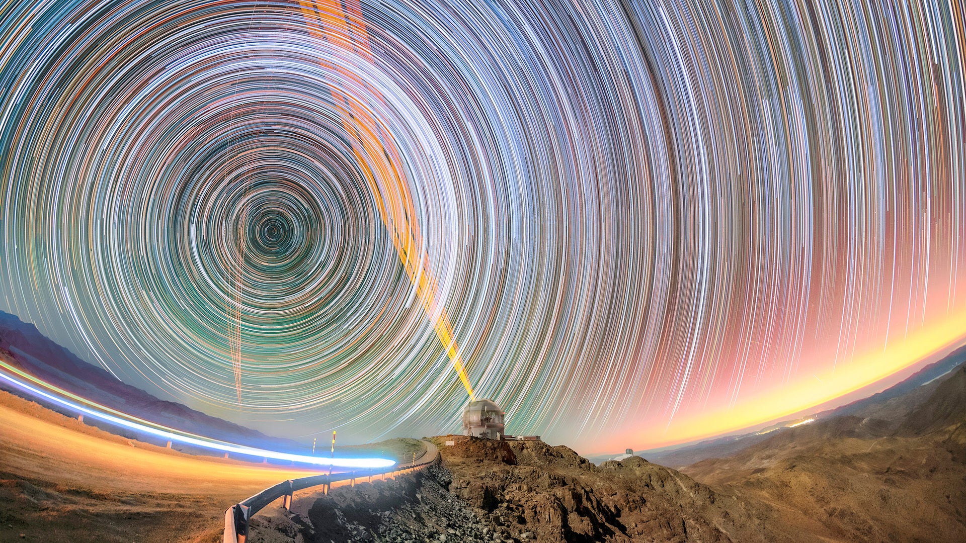 Langzeitbelichtung eines nächtlichen Himmels mit kreisförmigen Sternspuren über einem Bergobservatorium in einer kargen Landschaft.