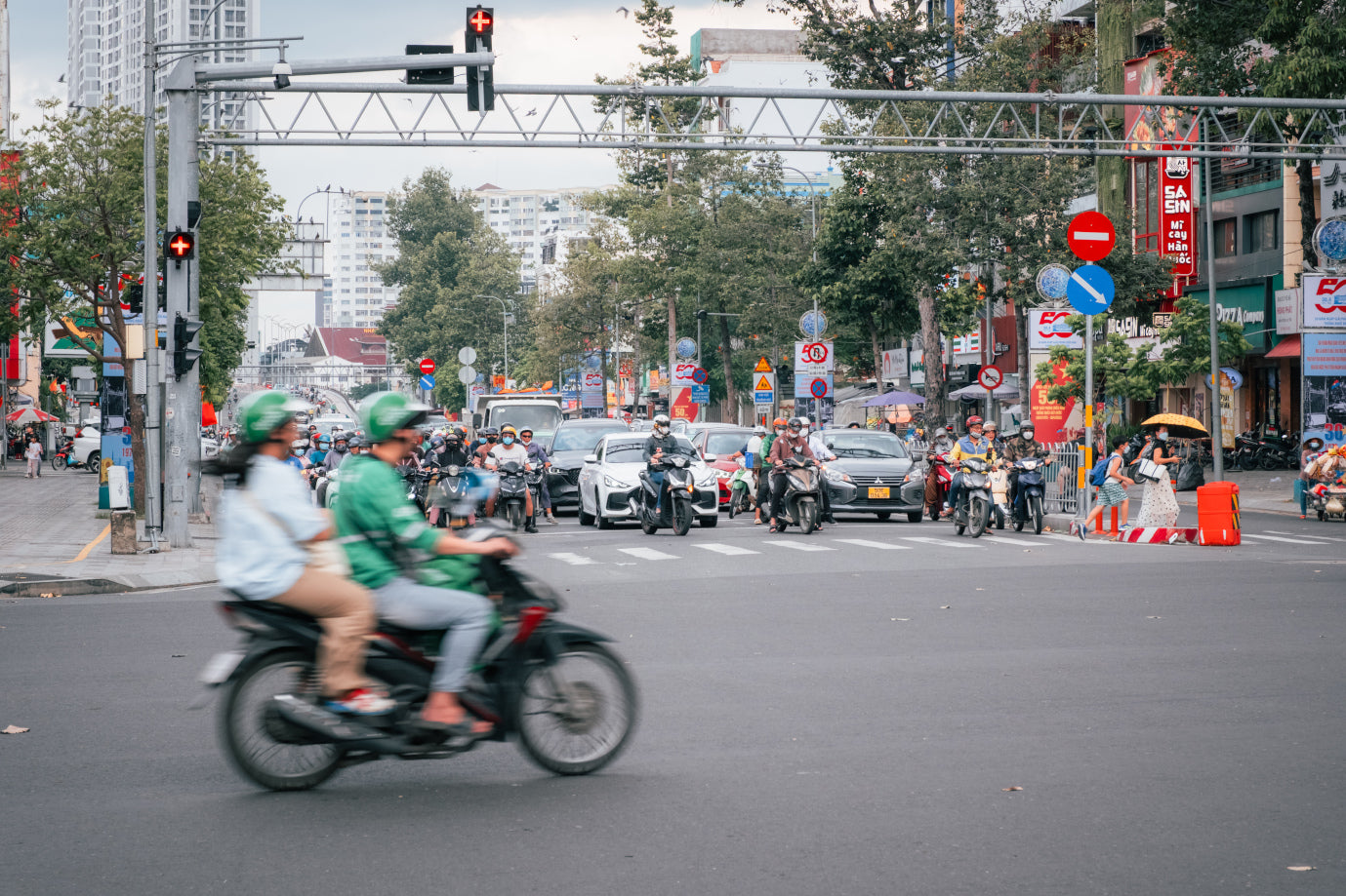 Belebte innerstädtische Kreuzung mit zahlreichen Motorrollern, Autos und wartenden Fußgänger:innen bei Rotlicht.