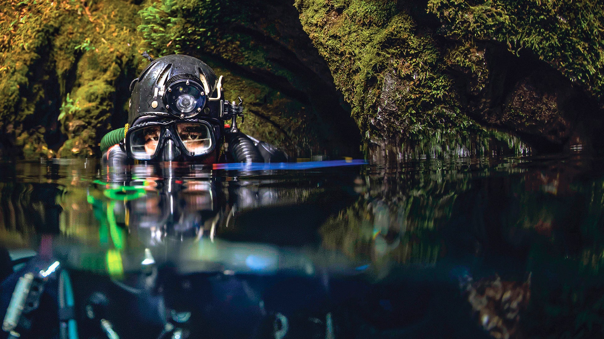 Höhlentaucher mit Tauchmaske und Stirnlampe taucht in dunklem Wasser nahe bewachsener Felswand auf, nur Kopf und Augen sind sichtbar.