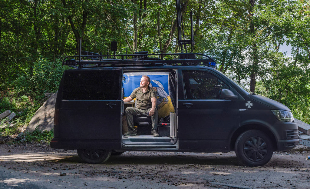 Mann sitzt im geöffneten schwarzen Transporter mit Antennen auf dem Dach, geparkt im Freien, genutzt als mobiles Kommando- oder Einsatzfahrzeug.