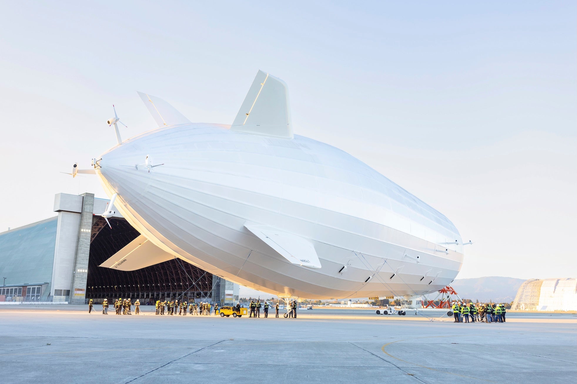 Ein großes weißes Luftschiff wird von vielen Menschen aus einem Hangar gezogen, während es für den Start vorbereitet wird.