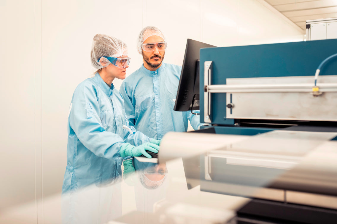 Zwei Wissenschaftler:innen in blauen Reinraumanzügen arbeiten an einer Hightech-Maschine und prüfen Daten auf einem Monitor.