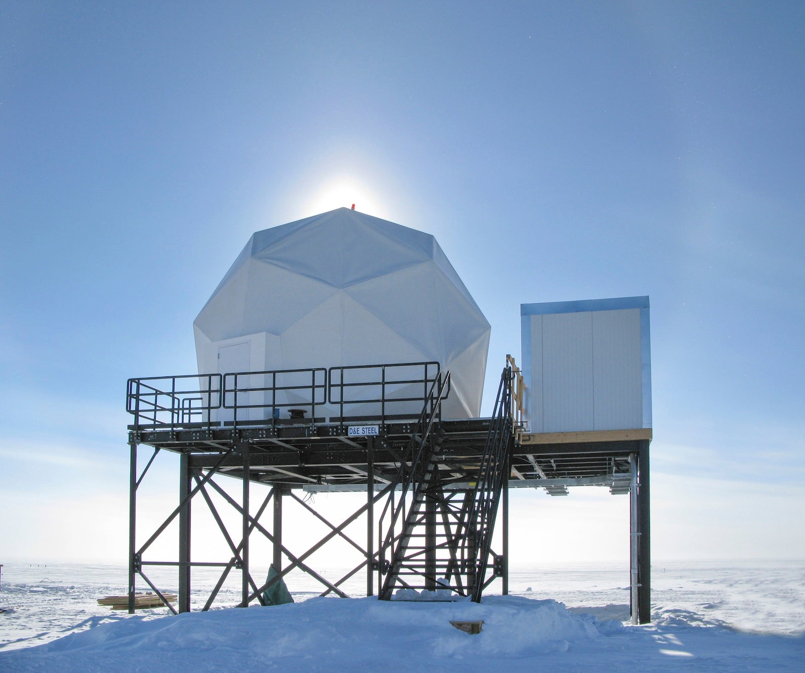 Weiße Kuppelstruktur einer Satelliten-Bodenstation auf Stahlkonstruktion in verschneiter Landschaft bei klarem Himmel.