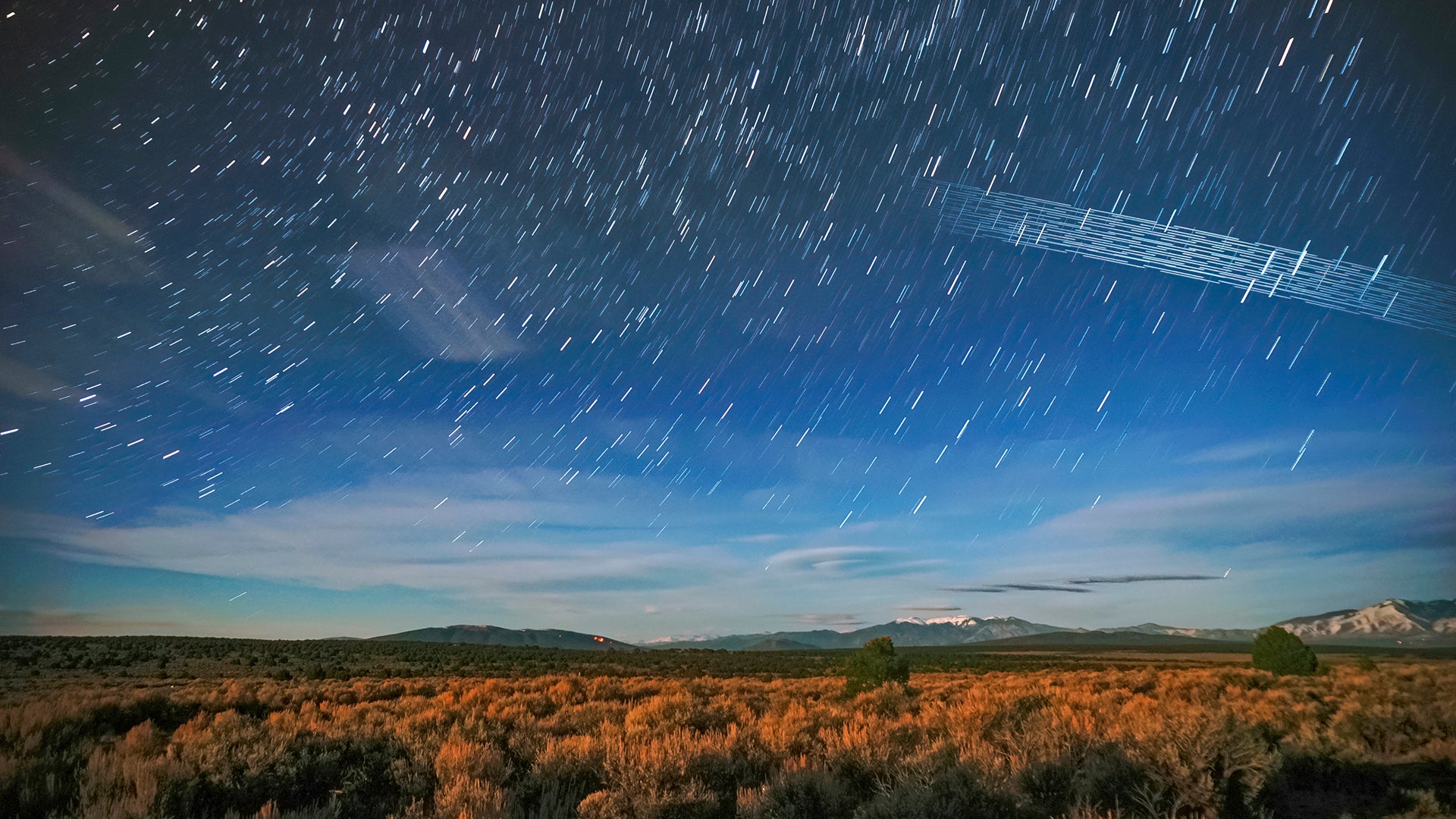 Langzeitbelichtung des Nachthimmels über einer Landschaft mit Starlink-Satelliten-Spuren und leuchtenden Sternen.