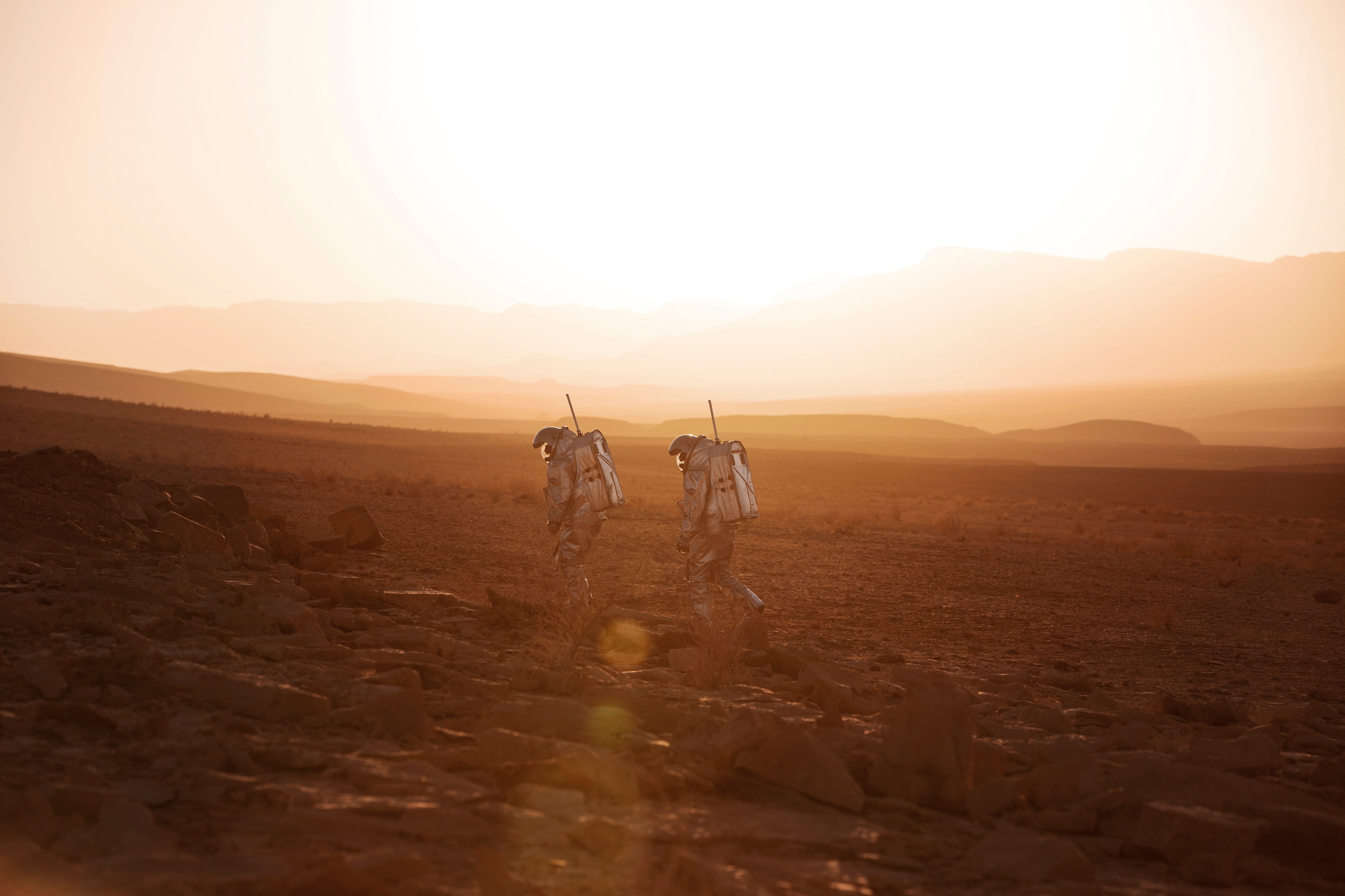 Zwei Astronaut:innen in silbernen Raumanzügen gehen durch eine karge Wüstenlandschaft im Sonnenuntergang. Szene erinnert an eine Mars-Simulation oder Weltraummission