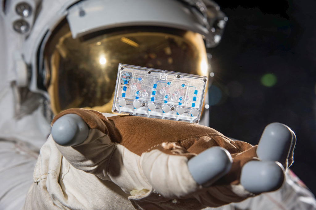 Astronaut im Raumanzug hält einen Tissue-Chip für biomedizinische Forschung im All in seiner Handschuhhand vor der Kamera.
