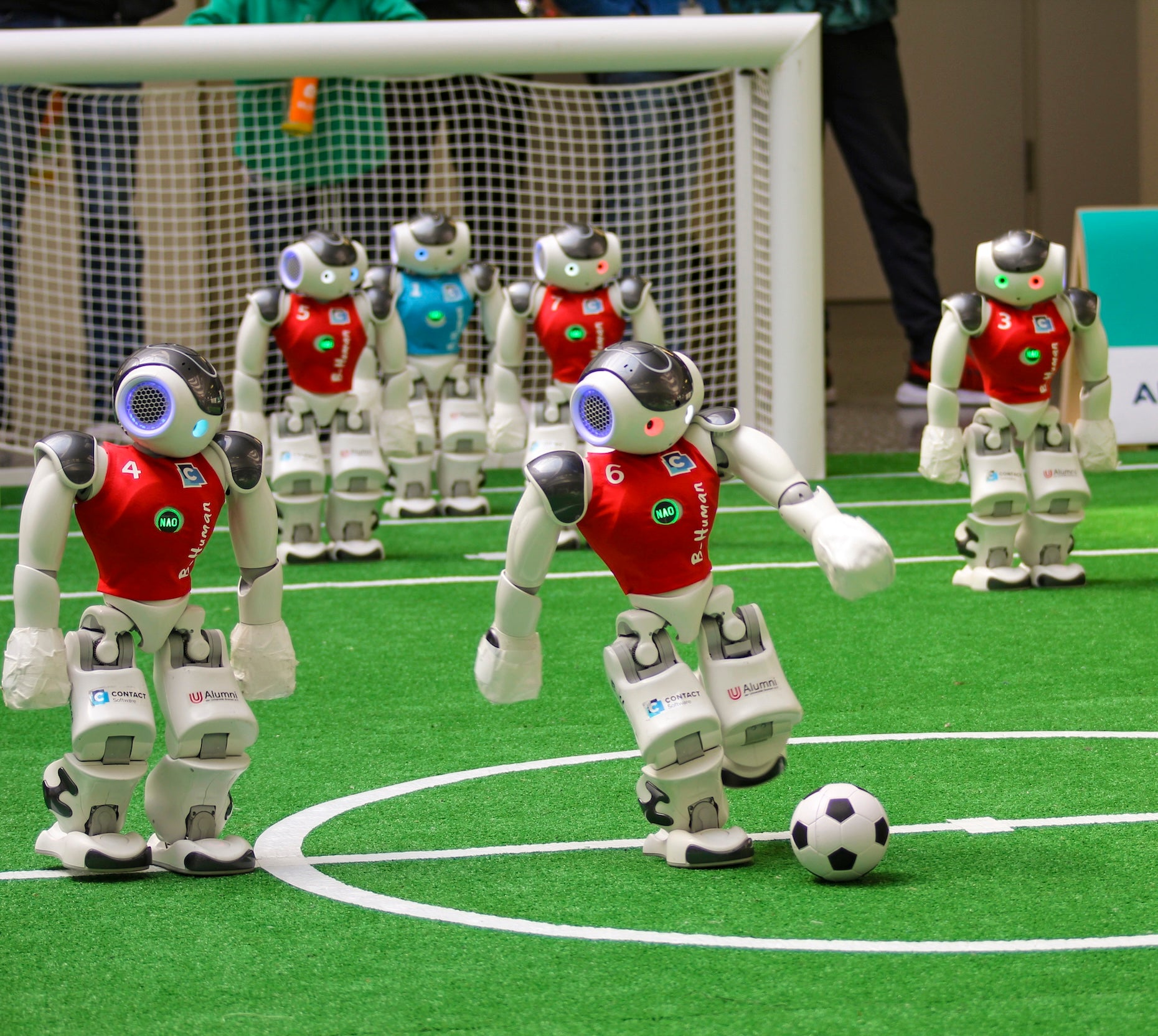 Mehrere humanoide Roboter im roten Trikot spielen Fußball auf einem grünen Kunstrasenfeld vor einem Tor.