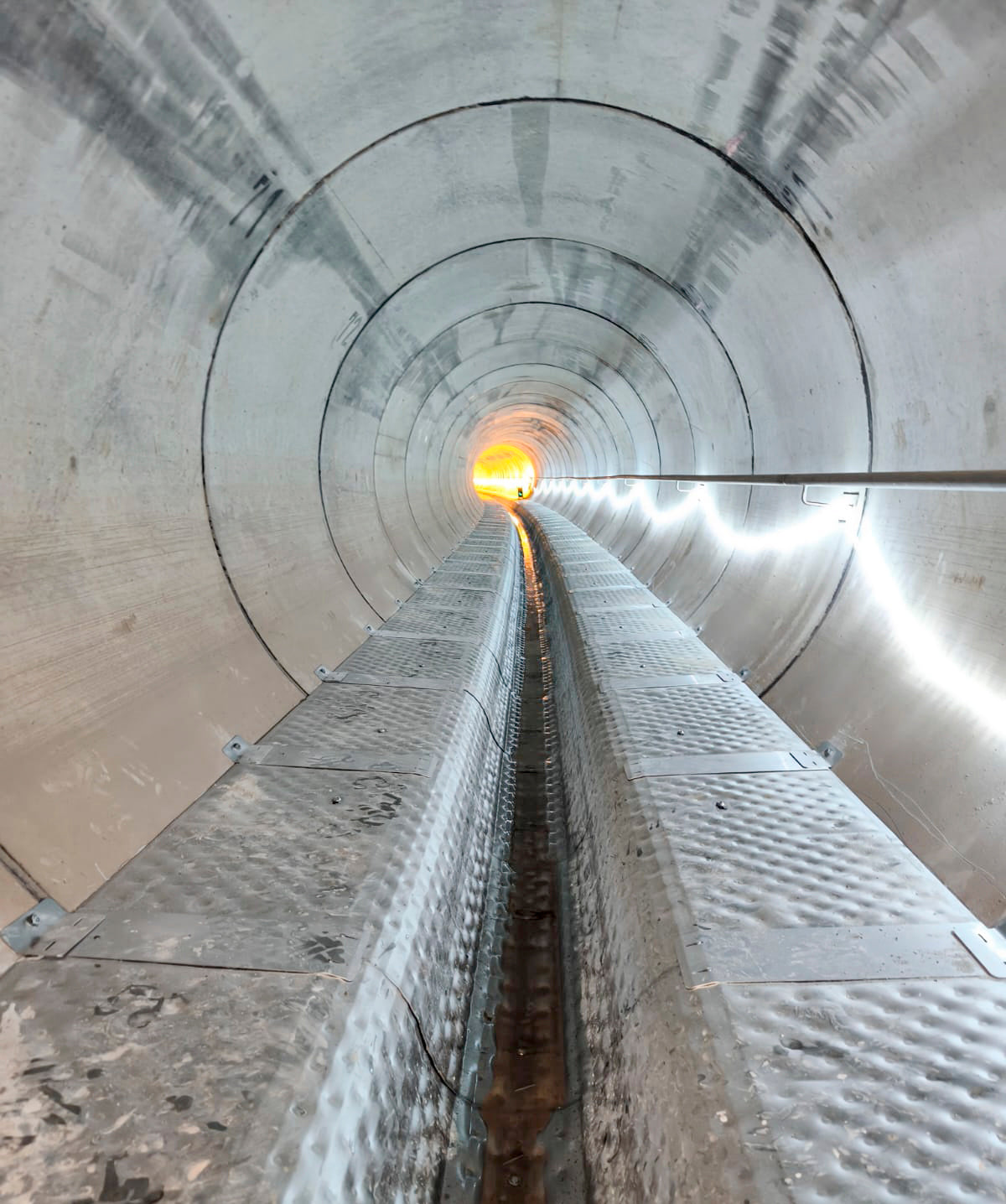 Blick durch einen beleuchteten, modernen Betontunnel mit metallischer Lauffläche bis zum hell erleuchteten Ende.