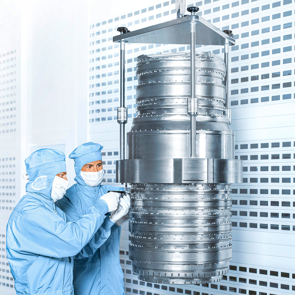 Zwei Ingenieur:innen in blauen Reinraumanzügen und Schutzmasken arbeiten an einem großen, silbernen Metallzylinder in einer Hightech-Fertigungsanlage. Darstellung für Präzisionstechnik und Halbleiterfertigung.